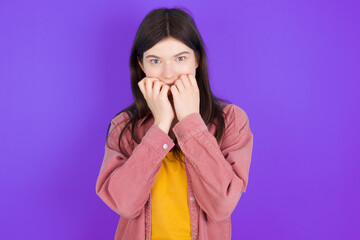 Anxiety - young beautiful Caucasian woman wearing casual clothes over purple wall covering his mouth with hands scared from something or someone bitting nails.