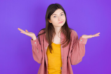 Puzzled and clueless young beautiful Caucasian woman wearing casual clothes over purple wall with arms out, shrugging shoulders, saying: who cares, so what, I don't know.