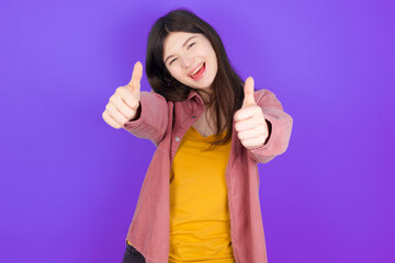 young beautiful Caucasian woman wearing casual clothes over purple wall making positive gesture with thumbs up smiling and happy for success. Looking at the camera, winner gesture.