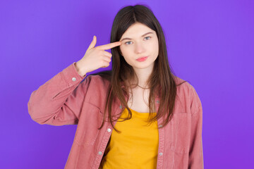 Unhappy young beautiful Caucasian woman wearing casual clothes over purple wall curves lips and makes suicide gesture,  shoots in temple with hand, tries kill himself.