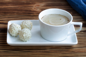 a cup of frothy latte stands on a square saucer on a wooden table with coconut sweets. Coffee with...