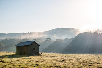 Sunrise morning silhouette of agricultural abandoned brick storage barn shed in autumn landscape farm field in rural countryside of West Virginia with fog mist sun sunrays by rolling hills