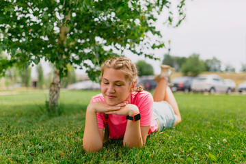 Naklejka na ściany i meble Caucasian girl with freckles and in a pink T-shirt lies on the grass. Girl student smiles and enjoys the beautiful summer weather.