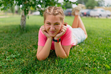 Caucasian girl with freckles and in a pink T-shirt lies on the grass. Girl student smiles and enjoys the beautiful summer weather.