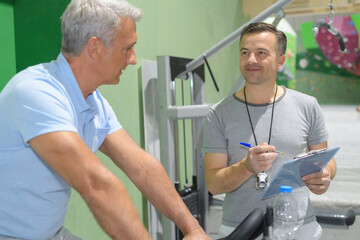 personal trainer holding clipboard with senior man using exercise equipment