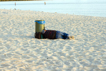 Samotny bezdomny człowiek śpiący na piaszczystej plaży w pobliżu pojemnika na śmieci - obrazy, fototapety, plakaty