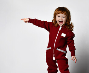 Cheerful smiling little red-haired toddler girl in warm jumpsuit pointing to a full-length standing sideways in front of the camera. Children's fashion and advertising of children's outerwear.