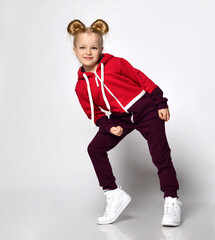 Little blonde curly beautiful girl in a red burgundy tracksuit and white sneakers leans on one knee with an elbow over a gray wall background. Stylish casual fashion for kids.