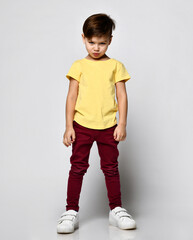 studio portrait of fashionable preschool little boy in yellow t-shirt, red denim pants, white sneakers, stands puffed out cheeks and lips, offended