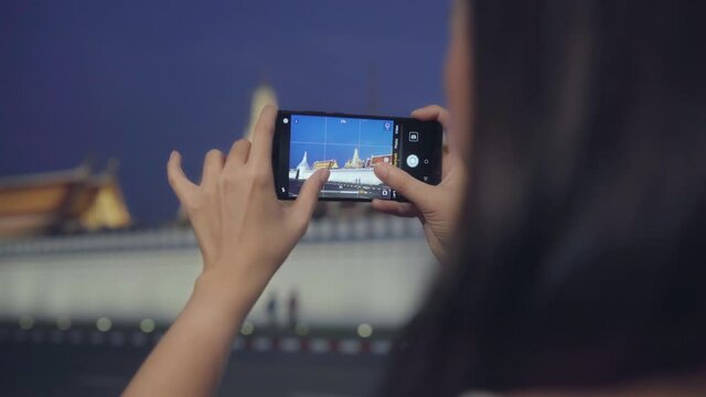 Happy young Asia girl tourist with casual style hold mobile phone relax cheerful take photo peaceful picture of temple in bangkok city at night. Lifestyle tourist travel holiday concept.