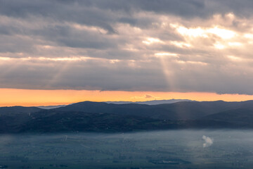 View from above of sunrays coming down on mist between hills and mountains in Umbria valley, Italy