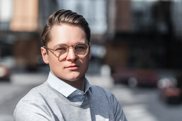 Fototapeta premium Portrait of young confident and serious business man with glasses, squinting from sun, looking camera, Stands against background of business center.