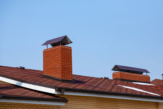 The Roof Of The Cottage To The Slopes, The Flashings, Chimney On Background Of Blue Sky. Brown Roof Of Bituminous Tiles.