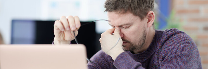 Tired man siting in front of laptop screen and holding glasses in hands