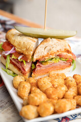 A spicy Italian sandwich with ham, salami, peppers, onions, tomatoes, pickle, cheese, and lettuce.  Served at an out door restaurant cafe.