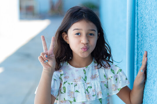 Uma criança, menina, brasileira dos cabelos pretos e lisos, fazendo biquinho encostada no muro pintado de azul. Fazendo sinal de "paz e amor" com os dedos.