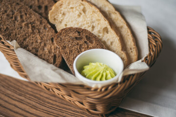 Wicker basket with black and white bread and bowl of homemade butter with herbs on white tablecloth on wooden table