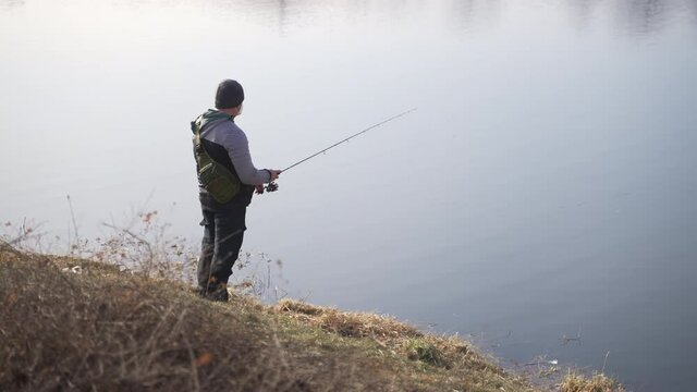 Footage of a man wearing a backpack using a fishing rod trying to catch fish from the river. Man waiting for fish. Hobby and Sport. Fish catching. River water in the background during the daytime