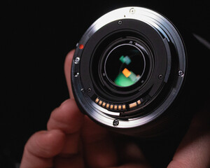 A hand holding a camera lens, showing the rear mounting plate.  Colors are reflected on the surface of the glass.  There is a solid, black background.
