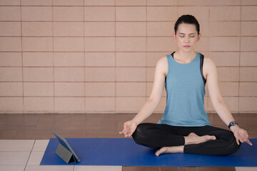 A middle-aged calm woman in relaxed sports outfits sits in a lotus position practicing yoga and meditation with relaxed eyes closed while listening to spiritual practices lesson on a tablet at home