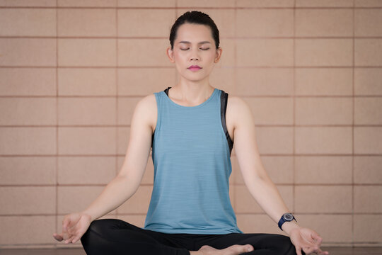 A middle-aged calm woman in relaxed sports outfits sits in a lotus position practicing yoga and meditation with relaxed eyes closed while listening to spiritual practices lesson on a tablet at home