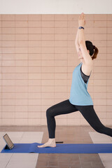 A middle-aged Asian woman in relaxed sports outfits doing an exercise workout training program follows an online workout lesson on a tablet at home during the COVID-19 pandemic and city lockdown