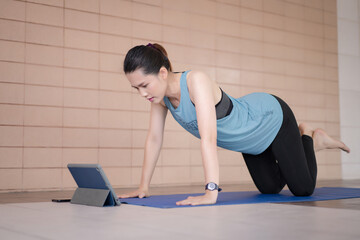 A middle-aged Asian woman in relaxed sports outfits doing an exercise workout training program follows an online workout lesson on a tablet at home during the COVID-19 pandemic and city lockdown