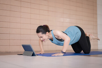 A middle-aged Asian woman in relaxed sports outfits doing an exercise workout training program follows an online workout lesson on a tablet at home during the COVID-19 pandemic and city lockdown