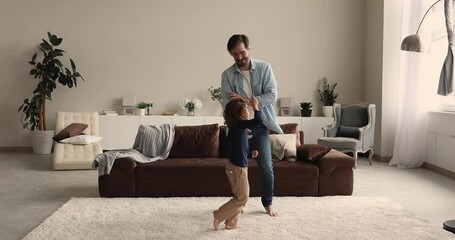 Happy energetic young father twisting in air joyful sincere small preschool child son at home, dancing to disco music. Emotional carefree family entertaining together on weekend leisure time.