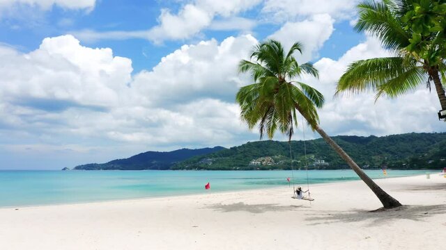 HD Phuket, Thailand. Tropical beach paradise with beach swing with girl in white shirt. Women relax on swing under coconut palm tree at beautiful tropical beach White sand.
