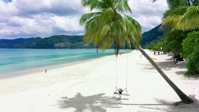 4K Phuket, Thailand. Tropical beach paradise with beach swing with girl in white shirt. Women relax on swing under coconut palm tree at beautiful tropical beach White sand.