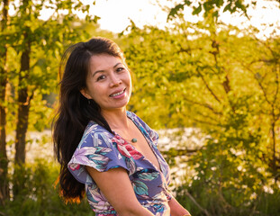 Fototapeta premium Portrait of smiling attractive Chinese woman with long black hair at sunset; woods in background