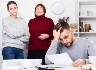 Portrait of distressed man with paperwork at home table with irritated family behind him