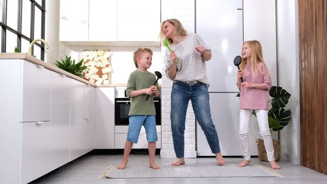 Excited mom or babysiter and two kids boy and girl jumping, dancing laughing in modern scandinavian house kitchen. Happy family mother daughter son having fun enjoy playing singing together at home.