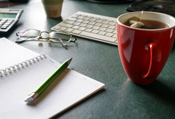 Home office. Notepad, keyboard, eyeglasses, pen and red cup with tea on desk.