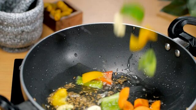 Closeup shot of adding colorful vegetables in a frying pan - cooking concept. Modern kitchen scene of a chef cooking mixed vegetables with cumin seeds in hot oil on an induction cooker
