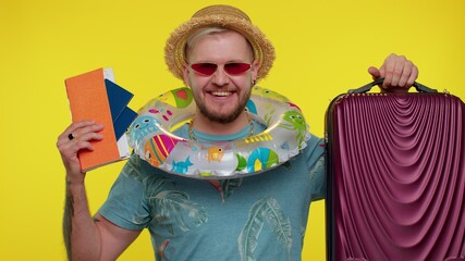 Fototapeta premium Traveler tourist bearded adult man celebrating, holding passport, tickets, luggage. Concept summer vacation, journey, trip to seaside. Isolated on studio yellow wall background. Guy air flight journey
