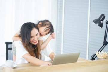 A Happy little Asian daughter and mother look at computer laptop together, have a cheerful expression in house on holiday
