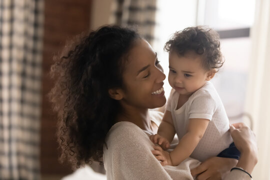 Smiling young African American mother play caress small toddler girl child at home. Happy biracial mom parent hug cuddle with little baby daughter kid, show love care. Parenthood concept.