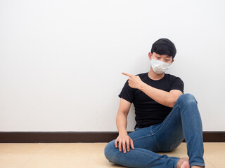 Sadly man wearing mask sit on the floor point finger left side to space white wall background