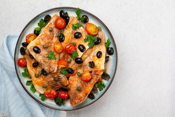 Perch fish fillets stew, italian style, with black olives, tomatoes and capers, with fresh parsley. On light blue plate. On light grey background. Top view.