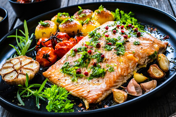 Roasted salmon steak with roast potatoes and vegetable salad served on black plate on wooden table
