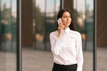 Portrait of a pretty Woman speaking on the phone wearing white business shirt. She is solving, negotiating daily tasks with her colleagues with glass walls in background.