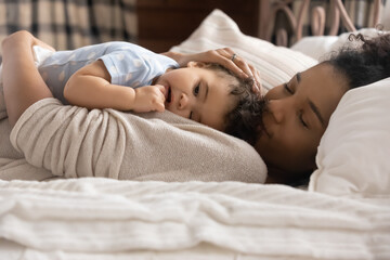 Happy loving young African American mom lying in bed caress cuddle little toddler daughter kid....
