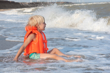 Light-haired boy in an orange inflatable vest sitting on seashore and waiting for big wave