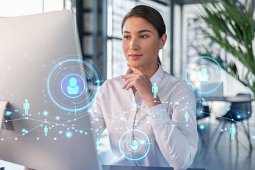 Attractive businesswoman in white shirt at workplace working with laptop to hire new employees for international business consulting. HR, social media hologram icons over office background