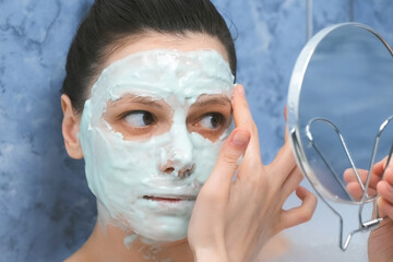 Portrait og young woman is applying alginate mask in bathr with foam looking at small mirror at home. Spa beauty treatment, anti-aging procedure face skin. Wellness, skincare and self love concept.