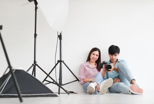 Portrait of two handsome man and beautiful woman wearing casual shirts, jeans, checking photos in camera while sitting on floor in photo studio with white background cutout and lighting equipment.