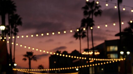 Defocused illuminated electric garland, palm trees silhouettes, Oceanside California USA. Ocean beach tropical pink sunset, pacific coast purple twilight sky. Los Angeles vibes. Bulb lights glowing.