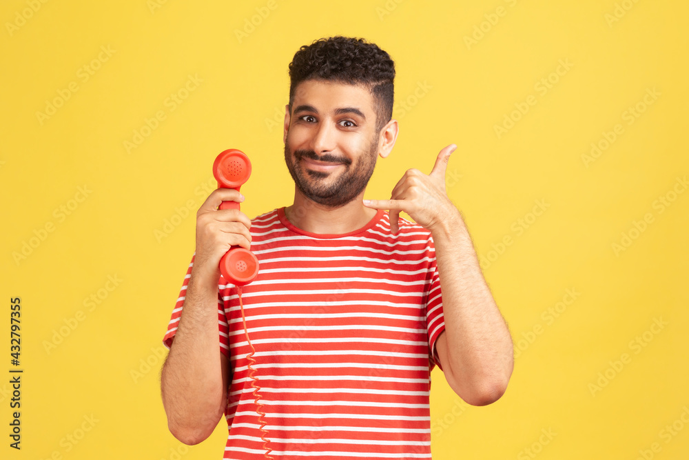 Wall mural Positive bearded man in striped t-shirt holding red headset of retro landline telephone and showing call me gesture, 24/7 call centre. Indoor studio shot isolated on yellow background